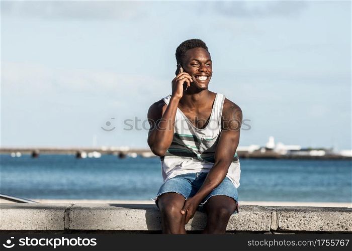 Optimistic ethnic male sitting on stone border on background of sea and having conversation on mobile phone while spending time on Lanzarote and looking away. Positive black man speaking on smartphone at seafront in summer