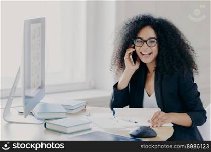 Optimistic female Afro American entrepreneur with curly hairstyle, holds mobile phone, enjoys telephone conversation with colleague, works on computer, poses at workplace with notepad and books