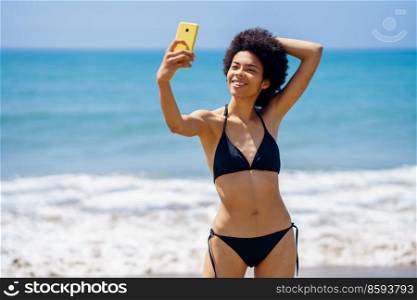 Optimistic fit African American fema≤trave≤r in swimwear taking self portrait on smartpho≠with hand behind head whi≤standing≠ar waving sea in∑mer. Content black woman in bikini taking selfie on beach