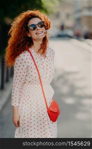 Optimistic foxy woman with glad expression, looks into distance, wears shades, long dress, strolls outdoor on street, enjoys nice summer day, has broad smile. People, lifestyle, rest and lifestyle