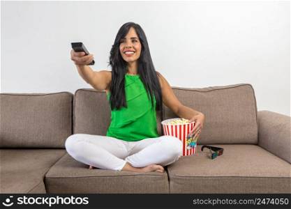 Optimistic Hispanic woman with bucket of popcorn smiling and switching channels on TV while sitting cross legged on couch during showtime at home. Cheerful female changing channels on TV