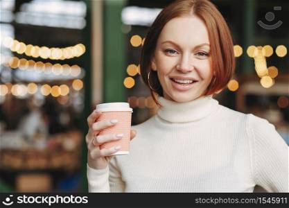 Optimistic lovely woman with dyed hair, satisfied expression, wears turtleneck jumper, holds takeaway coffee, poses in outdor restaurant, has nice beverage, being in high spirit, likes cappuccino