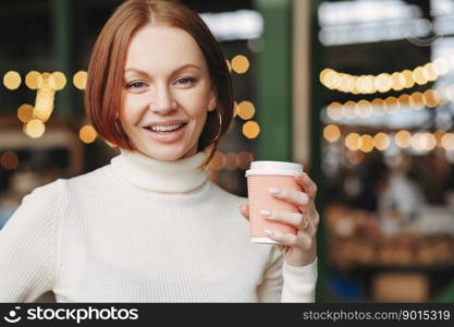 Optimistic lovely woman with dyed hair, satisfied expression, wears turtleneck jumper, holds takeaway coffee, poses in outdor restaurant, has nice beverage, being in high spirit, likes cappuccino