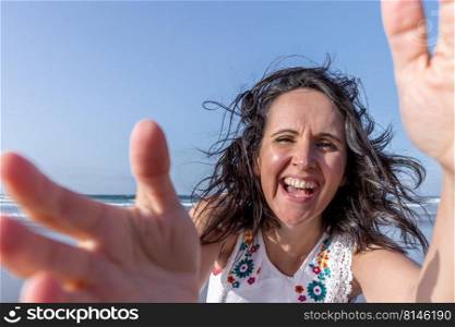 Optimistic middle aged woman with dark hair laughing and touching camera while having fun near sea against blue sky. Merry female traveler touching camera