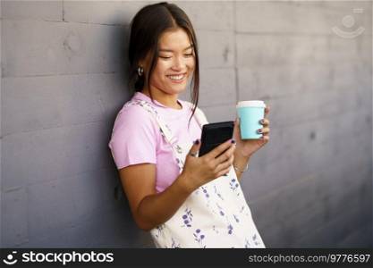 Optimistic young Asian female in stylish clothes with coffee to go smiling and watching video on cellphone while leaning on gray wall on city street. Cheerful Asian woman with coffee and smartphone leaning on wall