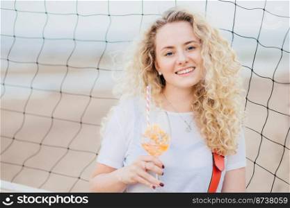 Optimistic young female model with curly light hair, dressed in casual white t shirt, drinks fresh cocktail, enjoys good rest at beach, stands near tennis net. People, beauty and rest concept