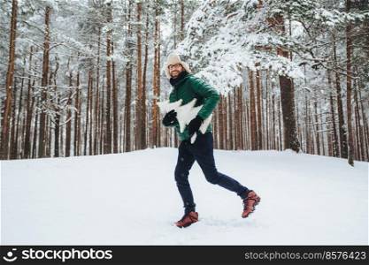 Optimistic young male dressed in warm winter clothes, has fun outdoor in winter forest, breath fresh air, glad to see much snow, holds atrficial fir tree in hands. People, season, weather concept