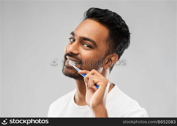 oral care, dental hygiene and people concept - smiling young indian man with toothbrush cleaning teeth over gray background. indian man with toothbrush cleaning teeth