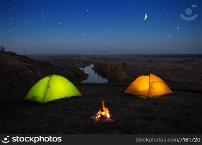 Orange and green tent by the river at night. Camping of two tents and a campfire on the river bank under a starry sky. Summer landscape. Concept of travel and solitude. Orange and green tent by the river at night