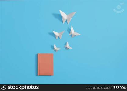 Orange hardcover notebook lying flat on a pastel blue background. Origami butterflies flying out of notebook into a blue sky. Copy space and room for text.