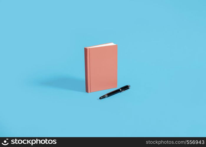 Orange hardcover notebook standing on a blue background with black pen. Center composition with space for copy and text.