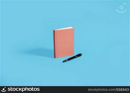 Orange hardcover notebook standing on a blue background with black pen. Center composition with space for copy and text.