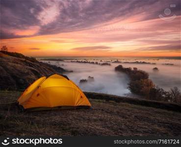 Orange illuminated from inside tent above river at sunset. Tent stands on hill above river covered with thick fog. Dramatic sunset. Summer landscape. The concept of privacy, travel and freedom.. Orange illuminated from inside tent above river at sunset