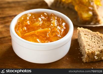 Orange jam in small bowl on wood with bread on the side, photographed with natural light (Selective Focus, Focus one third into the jam)