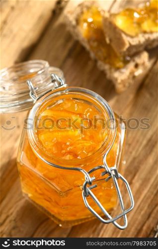 Orange jam in swing-top jar on wood with orange jam on bread in the back, photographed with natural light (Selective Focus, Focus on the first two orange peels on the jam)