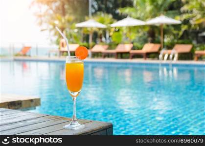 Orange juice glass on wooden table with swimming pool view backg. Orange juice with carrot slice in cocktail glass on wooden table at outdoor swimming pool, summer tropical holiday concept