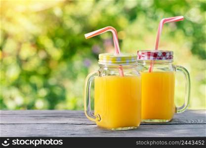 Orange juice in mason jars with the straws on a wooden table. Natural green background.. Orange juice in mason jars with the straws on wooden table