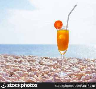 Orange juice with carrot slice in cocktail glass on white pebbly beach with seascape background, summer tropical holiday concept. Vintage filtered image.