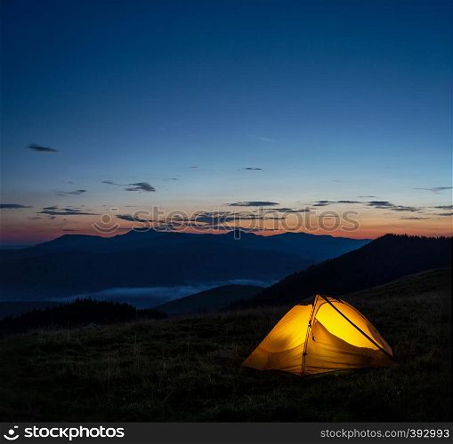 Orange lighted tent in the mountains under the evening sky. Summer landscape. The concept of freedom of solitude and travel. Orange lighted tent in mountains under evening sky