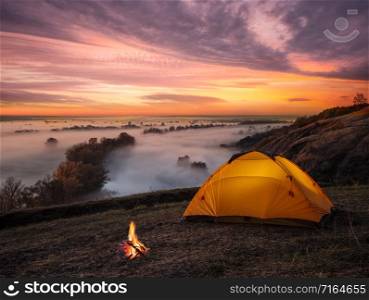 Orange lit inside the tent and a fire over the misty river at sunset. Dramatic sunset. Summer landscape. The concept of privacy, travel and freedom.. Orange lit inside tent and fire over misty river at sunset