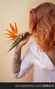 Orange strelitzia flower on the shoulder of a red-haired young girl with a tattoo around a beige background with copy space. Postcard. Strelitzia flower decorating the shoulder of a red-haired girl with a tattoo on a beige background with space for text. Creative layout