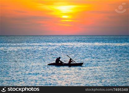 Orange sunset over blue sea and silhouette of kayak in rippled water