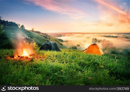 Orange tent and bonfire over the misty river. An orange tent and fire on hill above teak covered with a morning mist. Dramatic dawn sky. summer landscape. The concept of travel and freedom.. Orange tent and bonfire over the misty river