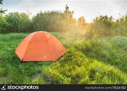 Orange tent camping at sunset in forest and green grass field and sun rays