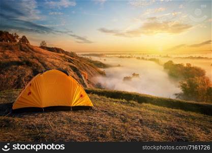 Orange tent in a canyon over a misty river at sunset. The concept of travel, freedom and privacy. Orange tent in canyon over misty river at sunset