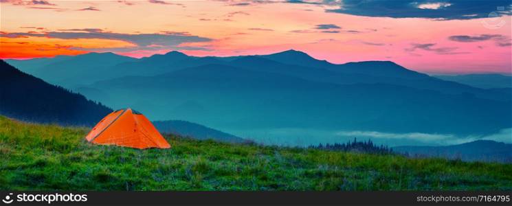 Orange tent in mountains at sunset. The chamber stands on a hill in the green grass. Against the background of silhouettes of mountains and dramatic sky. Solitude and travel wildlife concept. Orange tent in mountains at sunset