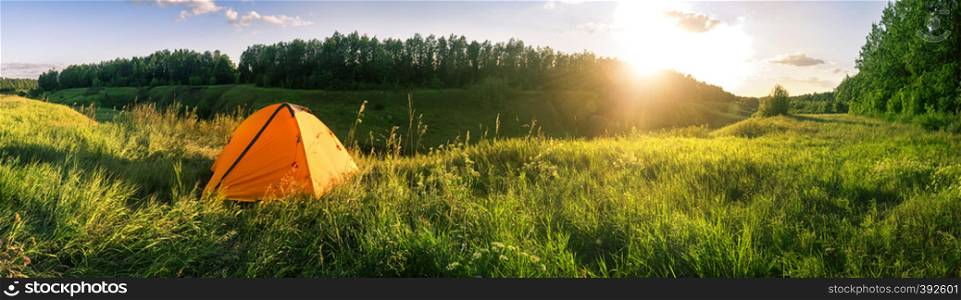 Orange tent in the field against the background of the forest. Sunset sky. Summer landscape. Panorama. Orange tent in field against background of forest
