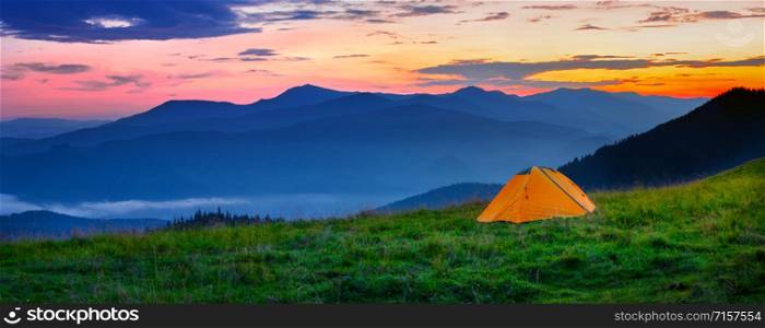 Orange tent in the mountains at sunset. The chamber stands on a hill in the green grass. Against the background of silhouettes of mountains and dramatic sky. Solitude and travel wildlife concept. Orange tent in the mountains at sunset