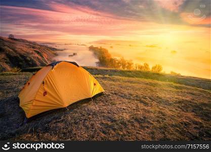 Orange tent on a hill above a foggy river under the bright sun. Dramatic sunset sky. Travel and privacy concept. Orange tent on hill above foggy river under bright sun