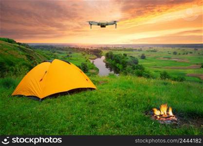 Orange tent on a hill above river under dramatic sky with quadcopter in air. Dawn over a camping and burning bonfire. Orange tent on hill above river under dramatic sky with quadcopter in air