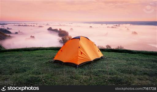 Orange tent on a hill above the misty river. Early morning. The concept of travel, freedom and privacy. Orange tent on hill above misty river