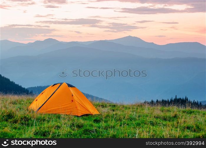 Orange tent on a meadow in the mountains under a pink dawn sky. Summer landscape. Orange tent on meadow in mountains under pink sky