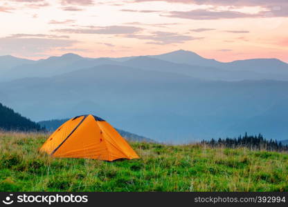 Orange tent on a meadow in the mountains under a pink dawn sky. Summer landscape. Orange tent on meadow in mountains under pink sky