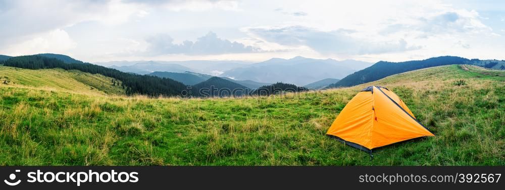 Orange tent on a meadow with green grass in the mountains. Cloudy sky. Summer landscape. Panorama.. Orange tent on a meadow with green grass in mountains