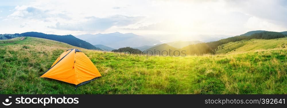 Orange tent on meadow with green grass in mountains under bright sun. Cloudy sky. Summer landscape. Panorama.. Orange tent on meadow with green grass in mountains under bright