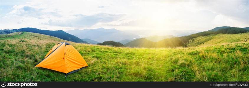 Orange tent on meadow with green grass in mountains under bright sun. Cloudy sky. Summer landscape. Panorama.. Orange tent on meadow with green grass in mountains under bright