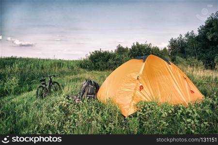Orange tent with backpack and bicycle on green meadow. Stylization for vintage. Toning. Orange tent with backpack and bicycle on green meadow