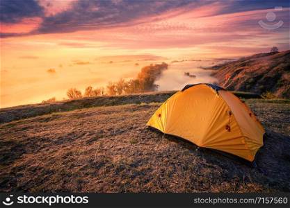 Orange tourist tent on a hill above a foggy river. Dramatic sunset sky. Travel and privacy concept. Orange tourist tent on hill above foggy river