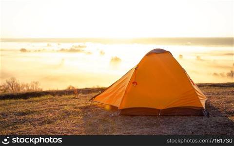 Orange tourist tent on a hill above the river covered with thick fog. Early morning. Summer landscape. The concept of travel, freedom and privacy. Orange tourist tent on hill above river covered with thick fog