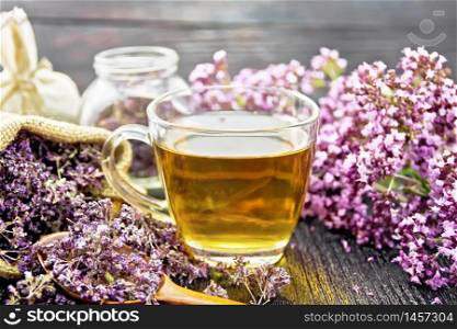Oregano herbal tea in a glass cup, fresh flowers, dried marjoram flowers in a bag, jar and spoon on wooden board background