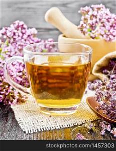 Oregano herbal tea in a glass cup on burlap, fresh flowers in mortar and on the table, dried marjoram flowers in a bag and spoon on wooden board background