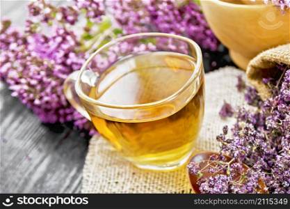Oregano herbal tea in a glass cup on burlap napkin, dried marjoram flowers in bag and spoon, fresh flowers in a mortar and on table against dark wooden board