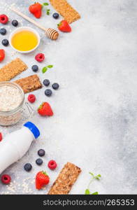 Organic cereal granola bar with berries with honey spoon and jar of oats and bottle of milk drink on stone background. Top view. Strawberry, raspberry and blueberry with almond nuts.