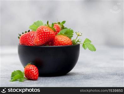 Organic fresh raw strawberries with leaf in black ceramic bowl on light table background.