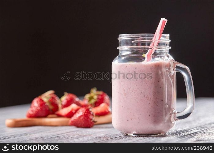 Organic milky strawberry smoothie in a glass with a straw on a gray wooden table around a dark background. Healthy dessert. Homemade strawberry milk shake in a jar with a straw on a wooden table around a black background. Organic vitamin dessert