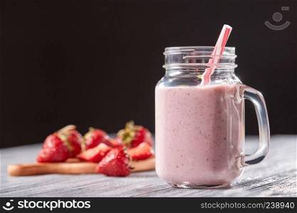 Organic milky strawberry smoothie in a glass with a straw on a gray wooden table around a dark background. Healthy dessert. Homemade strawberry milk shake in a jar with a straw on a wooden table around a black background. Organic vitamin dessert